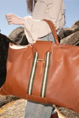 Bolso de viaje grande de cuero color marrón con cierre superior y correa con hebilla decorativa con rayas verdes y blancas.