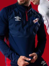 Buzo azul marino con detalles en rojo, cierre parcial con cremallera roja, cuello alto, logo de Umbro bordado en blanco y escudo del Club Nacional de Football bordado en el pecho.