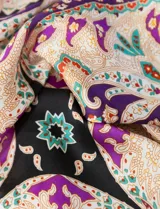 Bandana cuadrada de satén con estampado de arabescos en tonos violeta, naranja, celeste y blanco, con borde negro.
