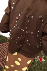 Chaqueta marrón de algodón con botones metálicos, cuello clásico, bolsillos frontales y apliques de tachas y botones formando una flor en la espalda.