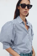Camisa de algodón a rayas verticales azules y blancas, con cuello solapa y mangas cortas abullonadas. Cierre frontal con botones.