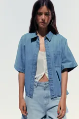 Camisa de denim celeste con cuello solapa, manga corta, bolsillo de parche en el pecho, bajo con aberturas laterales y cierre frontal con botones.