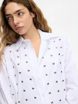 Camisa blanca de algodón con cuello camisero, cierre frontal con botones y tachas plateadas en el frente.