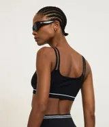 Top deportivo negro sin costuras, con escote asimétrico y doble tirante en los hombros. Presenta una delgada línea blanca horizontal en la banda inferior.