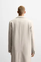 Abrigo recto color beige confeccionado en tejido con lana. Tiene cuello con solapas de muesca, manga larga con puños abotonados, bolsillos de vivo en la cadera, bolsillo interior, abertura en el centro de la espalda y cierre frontal con botonadura.