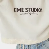 Sweater de punto grueso color blanco hueso, con cuello redondo, corte cuadrado y ligeramente corto. Presenta un gráfico frontal con el texto "EME STUDIOS curated by ben ice" en color marrón. Tiene puños ajustados.