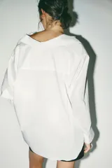 Camisa blanca amplia de cuello solapa y manga larga. Tiene un bolsillo de plastrón en el pecho, bajo asimétrico y cierre frontal con botones.