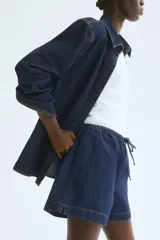 Short de denim azul oscuro, de corte holgado, con cintura elastizada y cordón de ajuste. Cuenta con bolsillos laterales y aberturas laterales en el ruedo.