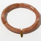 Pulsera rígida de color rosa con vetas blancas, compuesta por segmentos tubulares unidos por anillos dorados. Incluye un dije dorado en forma de gota.