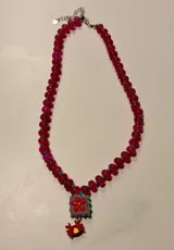 Collar de piedras naturales color fucsia, con pequeñas cuentas turquesas intercaladas. Incluye un dije cuadrado con una flor roja sobre fondo lila y un dije colgante en forma de cangrejo rojo.