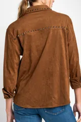Camisa marrón de gamuza vegana con cuello clásico, cierre frontal con botones y mangas largas con puños abotonados. Presenta detalles de tachas metálicas en cuello y charreteras.