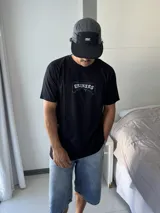 Remera negra de corte holgado con estampado gráfico en la espalda que dice "TAS LOCO" en letras rosas y celestes, con una ilustración de una ola y el texto "SUNNY WITH A CHANCE OF STOKE".