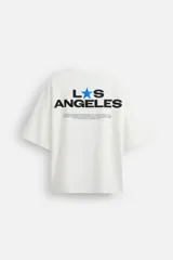 Camiseta blanca de corte holgado, cuello redondo y manga corta. Presenta un pequeño estampado de una estrella azul y un texto en negro en el frente.