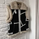 Chaleco negro de cuero sintético con interior de lana color crema, cierre frontal con cremallera y detalles plateados.