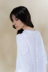 Blusa de lino blanca con escote a la base y terminación de voladito al bies desflecado. Delantera con alforzas verticales y mangas románticas tres cuartos, con detalle de puntilla. Puños con alforzas y voladitos que suman un toque femenino. Espalda abotonada.
