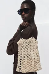 Bolso tipo shopper de tamaño pequeño, tejido en crochet color beige claro con patrón calado. Incluye asas de hombro de cuero sintético marrón oscuro y una bolsa interior con cierre de cremallera.