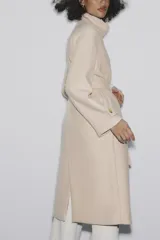 Abrigo largo color beige confeccionado en hilatura con lana. Tiene cuello subido, manga larga con trabillas, bolsillos laterales y cinturón ajustable con lazada. El cierre frontal es cruzado con botones.