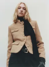 Chaqueta corta color beige de tweed con cuello mao, cierre frontal con botones y cuatro bolsillos delanteros con solapa.