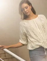 Blusa color crema de viscosa con cuello redondo y abertura central con botones. Tiene mangas 3/4 con puños fruncidos, detalles calados y bordados en mangas, delantera y espalda. La cintura tiene un lazo de ajuste.
