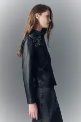 Camisa de símil cuero negra, de calce entallado, con cuello camisero y botones a presión con logo grabado. Presenta bordados florales con lentejuelas en los hombros y sobre los bolsillos frontales.