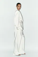 Blazer blanca de tejido satinado, con cuello solapa y escote pico, manga larga con hombreras, bolsillos con solapa en delantero y falso bolsillo en pecho, bajo con abertura en espalda y cierre frontal con botón.