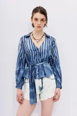 Camisa celeste con rayas verticales blancas, de corte holgado y mangas largas abullonadas con puños elásticos. Tiene cuello camisero, cierre frontal con botones y lazo en la cintura.