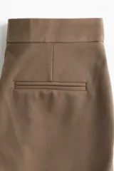 Pantalón de vestir color marrón claro, de tiro alto y corte bootcut. Presenta bolsillos laterales y traseros ribeteados, cierre frontal con botón y pliegues delanteros y traseros.