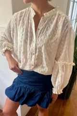 Camisa blanca con bordado de flores, cuello mao con volados, cierre frontal con botones de nácar y mangas largas abullonadas con puños elásticos con volados.