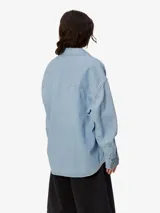 Camisa de denim azul de Carhartt WIP, de corte holgado, manga larga, cuello clásico y cierre frontal con botones. Tiene un bolsillo en el pecho con etiqueta con el logo de la marca.