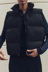 Chaleco blanco acolchado con cuello alto y cierre frontal con cremallera. Tiene bolsillos delanteros con cierre de cremallera oculta y bajo ajustable con cordones elásticos laterales.