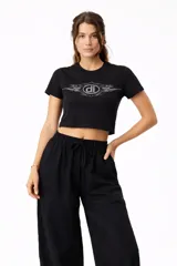 Remera negra de manga corta, corte ajustado y tiro corto (crop top), con un estampado frontal de estilo tribal o tribal-flameado en color gris claro que enmarca un logo circular con las letras 'dl'.