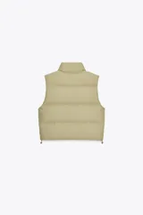 Chaleco blanco acolchado con cuello alto y cierre frontal con cremallera. Tiene bolsillos delanteros con cierre de cremallera oculta y bajo ajustable con cordones elásticos laterales.