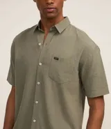 Camisa de hombre de corte relajado, color crudo, con cuello inglés, mangas cortas, abotonado delantero, bolsillo en el pecho y etiqueta de la marca.