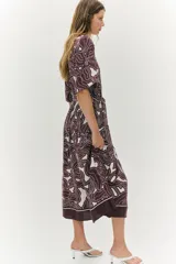 Vestido midi con cuello camisero, botonadura frontal y mangas cortas. Presenta un estampado abstracto en tonos marrón oscuro y blanco, elástico en la cintura y cinturón para atar.