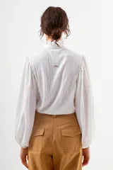 Camisa blanca de manga larga con cuello alto fruncido y puños ajustados. Presenta un corte holgado y tela con textura ligera.