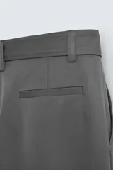 Pantalón negro de corte amplio con cinturón de tela con hebilla metálica. Presenta pliegues frontales, bolsillos laterales y traseros. Cierre frontal con cremallera y botón.