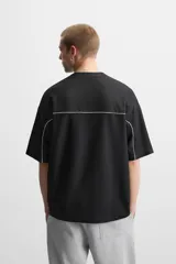 Camiseta negra de corte boxy fit, cuello redondo y manga corta. Presenta logos estampados en blanco en el frente y detalles en verde en los laterales.