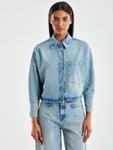 Chaqueta camisera de denim celeste, corta por delante y más larga por detrás, con cierre frontal con botones y bolsillo frontal aplicado en el pecho.