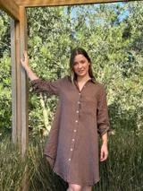 Vestido camisero marrón de lino, con cuello camisero, manga larga con botón para recoger, cierre frontal con botones de nácar, bolsillo de parche en el pecho y diseño asimétrico con ruedo más largo en la parte trasera.