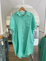Vestido camisero corto color verde agua, confeccionado en lino. Tiene cuello camisero, mangas cortas con dobladillo y un bolsillo de parche en el pecho. El corte es recto y holgado.