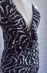 Vestido corto negro con lentejuelas plateadas dispuestas en un patrón ondulado o geométrico. Presenta un escote profundo en V pronunciado y tirantes finos tipo espagueti.