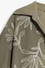 Camisa corta color verde oliva con bordado de hojas blancas, cuello solapa, escote pico y manga corta con vuelta.