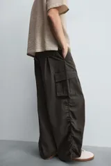 Pantalón cargo de corte ancho y relajado, color verde oscuro o negro, con cintura elástica, bolsillos laterales de parche y bolsillos traseros de vivo. Presenta pliegues verticales marcados en la parte delantera y cordones ajustables en los bajos.