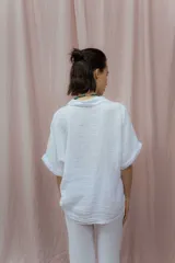 Camisa color crudo confeccionada en lino, de corte holgado, con cuello clásico, mangas cortas con dobladillo y cierre frontal con botones. Presenta detalle de pliegues verticales en la parte frontal.