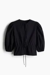 Blusa negra de algodón con textura de lunares, escote redondo con lazo, mangas tres cuartos abullonadas, botones frontales y cintura ajustable con cordón.
