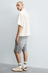 Bermuda de denim de algodón, color gris lavado, con corte relaxed fit y roturas visibles en la parte delantera de ambas piernas. Presenta cinco bolsillos y cierre frontal con cremallera y botón.