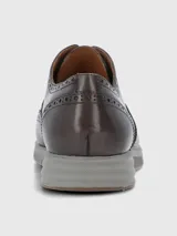 Zapato tipo Oxford o Brogue de cuero marrón oscuro, con puntera y laterales decorados con perforaciones (broguing). Posee cordones grises y una suela deportiva gruesa de goma color gris claro.