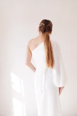 Vestido largo blanco de seda, con un hombro descubierto, manga larga abullonada y falda irregular con drapeado lateral. Incluye cinturón blanco con hebilla de alpaca y nácar.