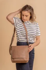 Bandolera de hombro color gris claro, con solapa frontal y correa ajustable. Presenta un diseño simple y práctico, ideal para uso diario.