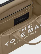 Bolso tote mediano color beige con estampado frontal del logo "The Tote Bag Marc Jacobs" en blanco. Tiene doble asa corta y correa larga extraíble de tela azul marino.